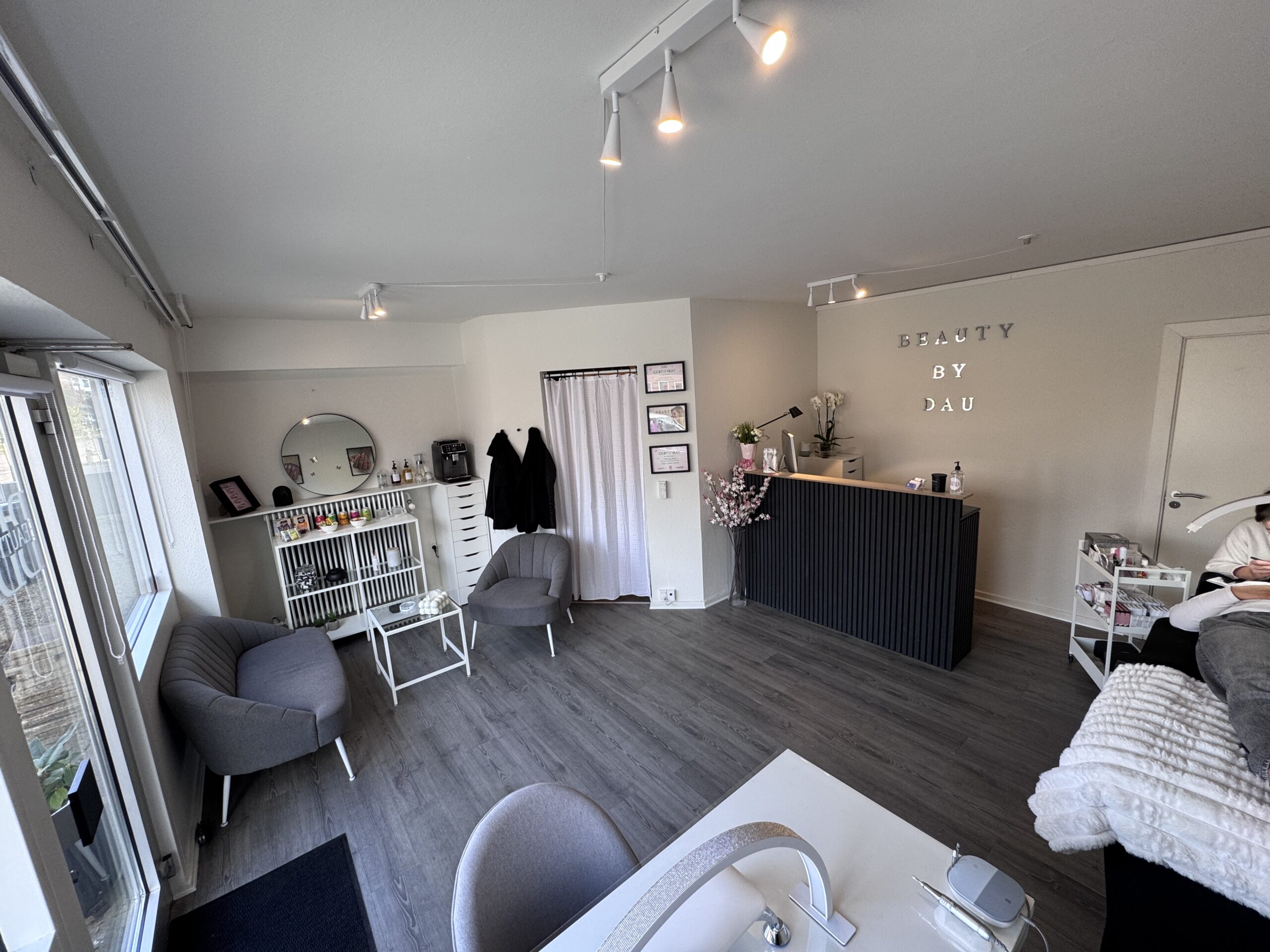 Indretning af BeautyByDau’s skønhedsklinik med lyse vægge, hyggelige stole, reception og produkter på hylder. Et indbydende og roligt behandlermiljø i stilrene omgivelser.