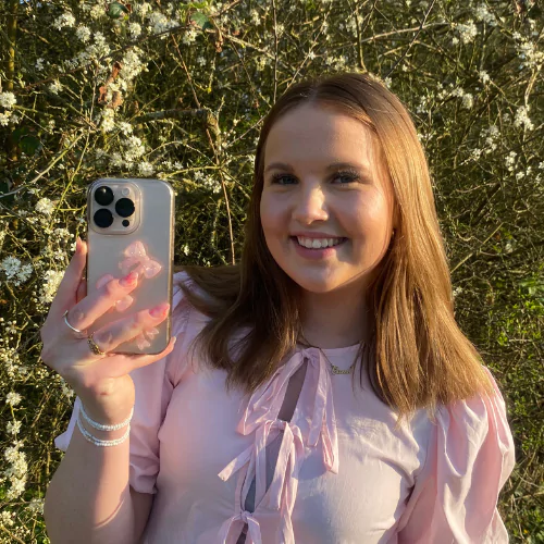 Camille, ung content creator hos BeautyByDau, smilende i lyserød bluse med telefon i hånden foran blomstrende busk.