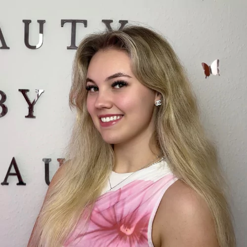 Emilie, klinikchef hos BeautyByDau, står foran logo-væg i hvid top med pink blomst og smiler til kameraet.
