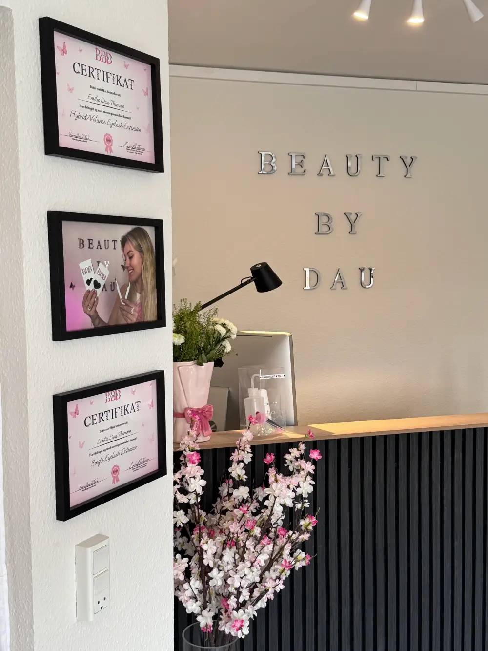 Receptionen hos BeautyByDau i Sønderborg med diplomer på væggen og elegant blomsterdekoration. Viser certificeringer i eyelash extensions og en professionel velkomststemning.